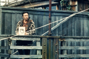 Alan Watts at his houseboat, smoking pipe, holding a book.